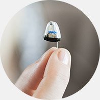[GEN] A hand holds a small, discreet Oticon hearing aid with a transparent casing, showcasing advanced technology. The soft-focus background emphasizes the device's compact design, ideal for personalised hearing care.