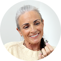 Una persona mayor sostiene un audífono discreto con diseño intraauricular en la mano derecha. Lleva un suéter beige y está en un entorno iluminado, posiblemente una clínica auditiva.