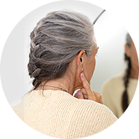 Una mujer con cabello canoso trenzado ajusta un audífono detrás de su oreja izquierda frente a un espejo. Lleva un suéter beige en un entorno luminoso y minimalista.