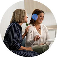 Dos mujeres sentadas conversan; una muestra interés tocando el brazo de la otra. Contexto acogedor en una clínica auditiva. Se resalta un audífono azul detrás de la oreja derecha.