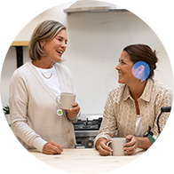 Dos mujeres conversan mientras sostienen tazas en una cocina iluminada; una de ellas lleva un audífono visible de color azul. Fondo incluye estantes y electrodomésticos modernos.