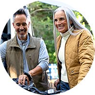 Two people, a man in a brown vest and a woman in a mustard quilted jacket, stand outdoors next to a bicycle, surrounded by greenery in a casual setting.
