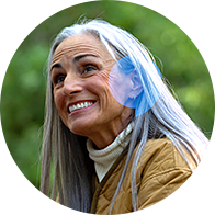 Una donna con lunghi capelli grigi indossa un apparecchio acustico discreto dietro l'orecchio sinistro. Si trova in un ambiente naturale, illuminato da luce soffusa tra alberi verdi.