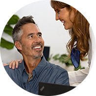 Een man zit aan een bureau met een laptop, terwijl een vrouw in een witte jas met een blauwe sjaal hem ondersteunend op de schouder aanraakt in een professionele, klinische omgeving.