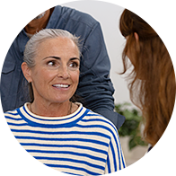 Una donna anziana con capelli grigi raccolti indossa una maglia a righe blu e bianche; è seduta in un contesto di consulenza, affiancata da due persone in un ambiente di un centro acustico.