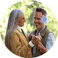 Una coppia anziana balla all’aperto tenendosi per mano; l’uomo indossa un apparecchio acustico sull’orecchio destro. Lo sfondo mostra alberi e luce naturale.