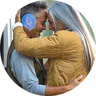 Una coppia abbracciata, con la donna dai capelli grigi e giacca gialla e l’uomo che indossa un apparecchio acustico sull’orecchio destro; ambiente esterno sfocato.