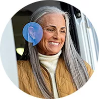 Una persona con capelli grigi indossa un apparecchio acustico poco visibile sull'orecchio sinistro. Indossa un cappotto marrone e un maglione bianco, affacciandosi da un veicolo.