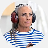 Une personne, portant un casque de protection auditive bicolore (rouge et noir), tient un objet dans un environnement lumineux. Le pull rayé blanc et bleu suggère une ambiance décontractée.