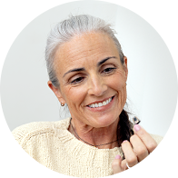 Uma pessoa idosa segura um pequeno aparelho auditivo em uma das mãos, demonstrando interesse ou uso. O fundo é claro, sugerindo o ambiente num centro auditivo.