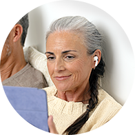 Uma mulher idosa, de cabelos grisalhos e trançados, usa um aparelho auditivo no ouvido direito enquanto segura um tablet em ambiente doméstico ao lado de um homem.