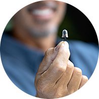 Een hand houdt een klein, discreet hoortoestel omhoog, terwijl een glimlachende man op de achtergrond zichtbaar is. De context suggereert een focus op gehoorzorg en innovatieve hoorhulpmiddelen.