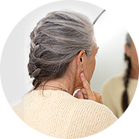 Una donna anziana con capelli grigi intrecciati tocca l'orecchio vicino a uno specchio, indossando un maglione beige in un ambiente luminoso.