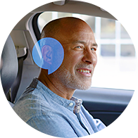 A person seated in a car wearing a blue shirt, with a digitally superimposed blue ear icon suggesting the usage of a hearing aid, emphasizing hearing care solutions in daily life.