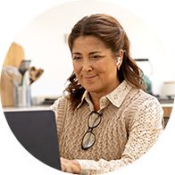 Una donna indossa un apparecchio acustico mentre utilizza un computer portatile in una cucina luminosa; occhiali da lettura pendono al collo e utensili da cucina sono sullo sfondo.