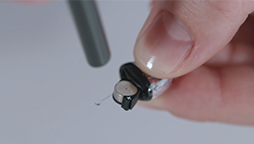 Een hand houdt een klein hoortoestel vast terwijl een hulpmiddel de batterijplaats inspecteert; de context is een verzorgde, neutrale achtergrond, typisch voor een hoorwinkel of audioloog.