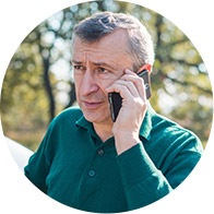 Un hombre mayor con camisa verde sostiene un teléfono móvil junto a su oído. Está al aire libre, rodeado de árboles en un entorno natural con luz diurna.