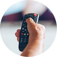 Una mano sostiene y usa un control remoto negro para cambiar de canal o ajustar funciones, en un ambiente borroso que sugiere un televisor encendido en un hogar.