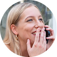 Une femme blonde porte des boucles d'oreilles dorées et tient un appareil noir près de son visage. À l'arrière-plan, une façade vitrée évoque un contexte urbain ou commercial. Aucun texte visible.