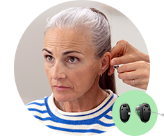 A person with gray hair is fitted with a hearing aid by a professional's hand in a clinical setting. A green icon displays a pair of black Oticon hearing aids nearby.