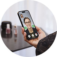 A person holds a smartphone displaying a video call, with blurred face visible onscreen. The background includes a table with nail polish bottles, and the setting appears indoors.