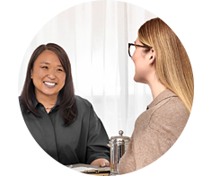 A woman with glasses consults a professional in a hearing clinic, seated at a table with documents and a small metallic device. The setting suggests a friendly hearing care appointment.