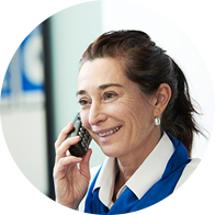 Una donna, con indosso una divisa blu e bianca, parla al telefono in un contesto professionale, probabilmente una clinica Audika specializzata in apparecchi acustici, assistenza uditiva e test dell’udito.