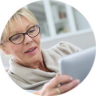 Een persoon houdt een tablet vast en kijkt ernaar, zittend in een lichte, huiselijke omgeving met een kast op de achtergrond. Geen zichtbare tekst of gehoorapparaten.