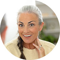 Une femme aux cheveux gris, portant un pull beige, touche son oreille, suggérant un problème auditif ou une écoute attentive.