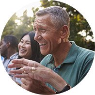 An older man wearing a discreet behind-the-ear hearing aid engages in conversation outdoors, surrounded by others in a relaxed, sunlit park setting.