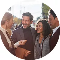 Quatro pessoas interagem em uma conversa ao ar livre, em um ambiente urbano ensolarado, com árvores e edifícios ao fundo. Os rostos estão desfocados, impossibilitando a identificação.