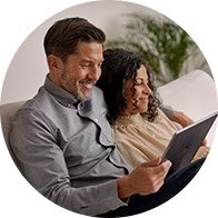 En voksen og et barn sidder tæt sammen i en sofa og kigger på en tablet. I baggrunden ses en diskret, lys stue med en grøn plante. Ingen tekst eller høreapparater er synlige.