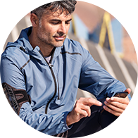 Un uomo in giacca sportiva blu tocca lo schermo di uno smartphone con un dito, seduto in un contesto esterno urbano con gradini sfocati sullo sfondo. 