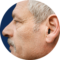 Orelha de uma pessoa idosa usando um aparelho auditivo discreto intra-auricular. O fundo é desfocado, destacando o aparelho e sugerindo um contexto relacionado à saúde auditiva ou centro auditivo.