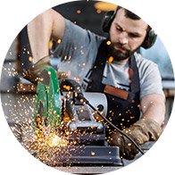 Een arbeider gebruikt een slijpmachine, waarbij vonken vliegen tijdens het slijpen van metaal. De werkomgeving is een werkplaats, en de persoon draagt gehoorbescherming en handschoenen. Geen tekst aanwezig.
