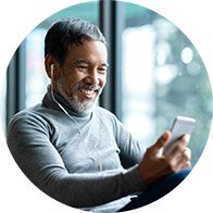 [GEN] A person wearing a gray turtleneck listens through wired earphones while holding a smartphone, seated in a modern, well-lit room with large windows overlooking blurred outdoor greenery.