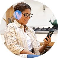 A person wearing a discreet hearing aid sits in a kitchen environment, holding and looking at a smartphone. The context highlights modern hearing solutions like those from hearing care experts.