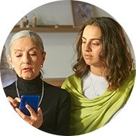 Duas pessoas em um ambiente interno, com rostos desfocados, visualizam juntas um celular azul. Ao fundo, prateleiras com pequenos objetos decorativos compõem o contexto acolhedor.