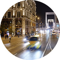 Een drukke stadsstraat bij nacht met felverlichte gebouwen links, passerende auto's en een verlichte brug op de achtergrond, die een levendige stedelijke sfeer creëert. Geen tekst aanwezig.