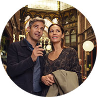 [GEN] A couple stands closely, one holding a small device resembling a hearing aid, within an ornately decorated indoor space featuring arched ceilings, intricate ironwork, and glowing spherical lamps. No visible text or brand is identifiable.