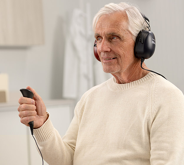 Un homme âgé porte un casque noir marqué 'DECIBEL' et tient un bouton-poussoir dans une main, lors d’un test auditif dans un cabinet calme et lumineux d’Audika.