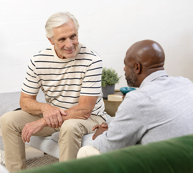 Un homme âgé en polo rayé discute avec un audiologiste dans un cadre chaleureux. Des plantes en pot et un canapé vert créent une ambiance accueillante, suggérant une consultation en clinique auditive.