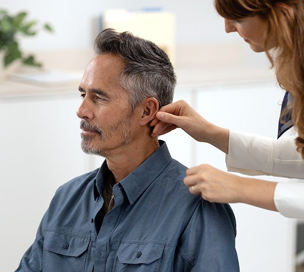 Un professionnel ajuste un appareil auditif sur l’oreille d’un homme assis dans un cabinet.