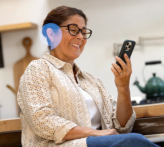 Woman with glasses wearing a hearing aid while using a smartphone in a kitchen setting.
