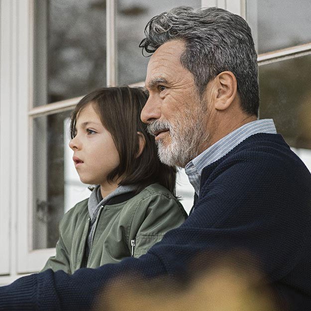 A middle-aged man with graying hair and a child sit closely together outdoors near a large-paneled window. The man wears a navy sweater; the child wears a green jacket. No visible hearing aids or text are present.