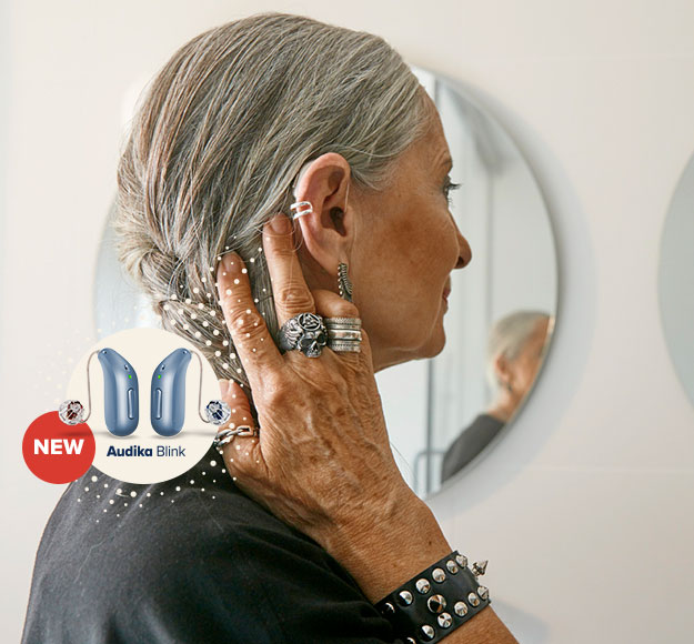 Una donna anziana con capelli grigi raccoglie i capelli vicino all'orecchio, indossando un apparecchio acustico. Sul lato sinistro appare il testo: 'NEW Audika Blink'. Sfondo: specchi circolari in un ambiente moderno.