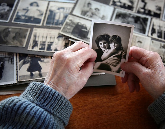 Deux mains d’une personne âgée tiennent une photo en noir et blanc, entourées de plusieurs photos anciennes posées sur une table en bois.