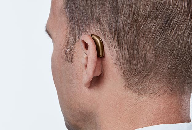 Un homme porte une aide auditive beige derrière l’oreille droite, soulignant une solution discrète et technologique pour améliorer l’audition. Fond neutre, typique d’un contexte de clinique auditive.