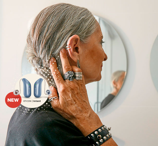 A senior woman with gray hair adjusts a discreet hearing aid behind her ear while standing in front of a circular mirror. The image highlights 'NEW Oticon Intent' hearing aids in a promotional overlay.