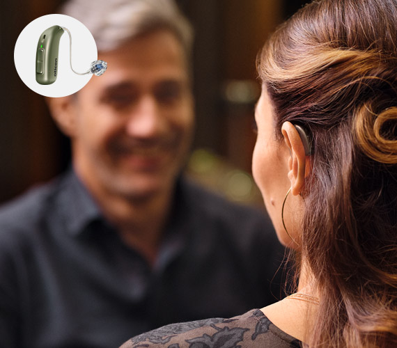 A woman wearing a discreet hearing aid engages in a conversation in a warmly lit environment. The inset showcases an Oticon hearing aid, highlighting advanced hearing care technology by Audika.